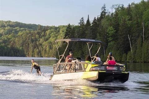 2024 Sea-Doo Switch Sport 21 on a scenic lake with wakeboarder and lush forest backdrop.