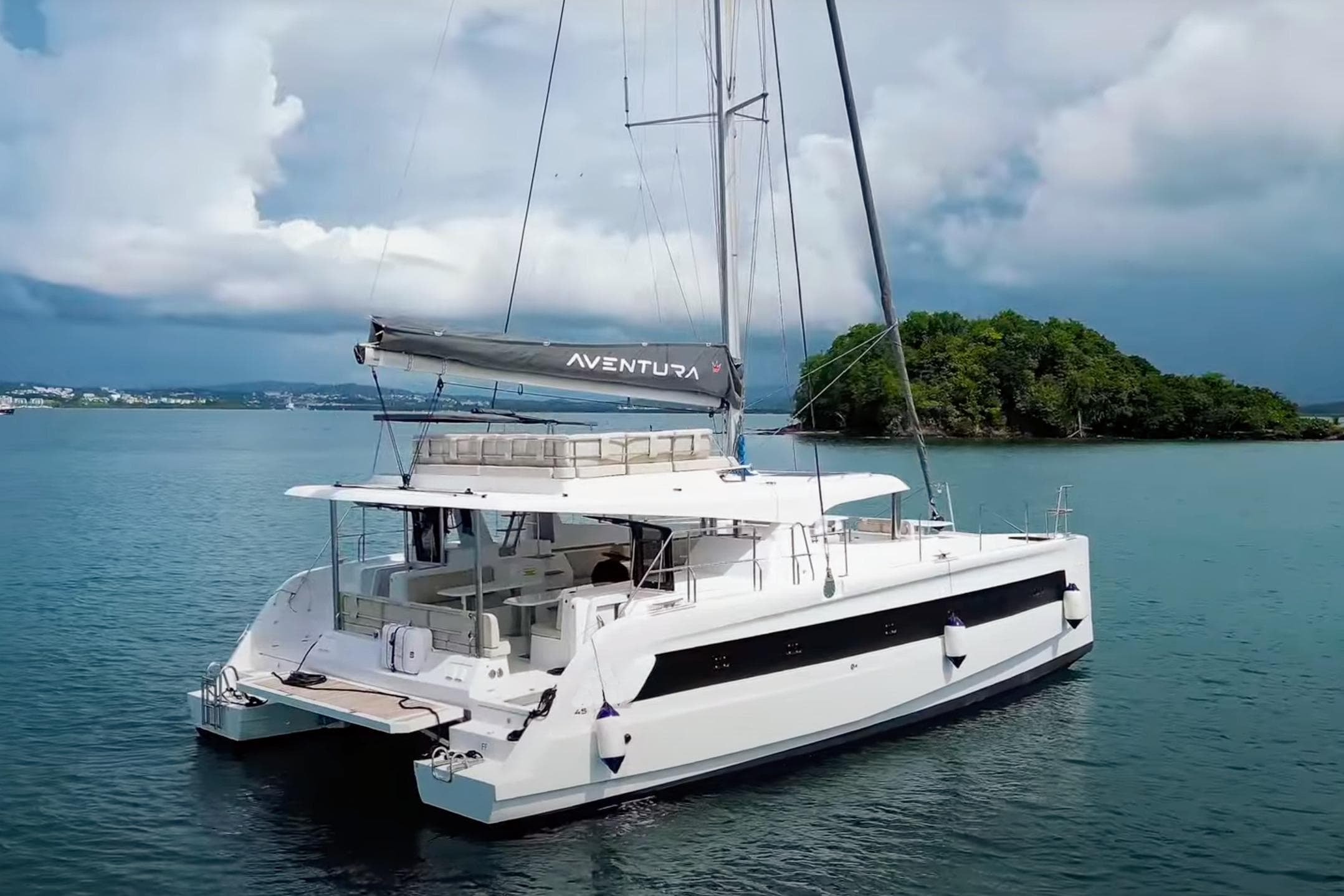 Aventura 45 catamaran sailing near an island, 2025 model, under cloudy skies.