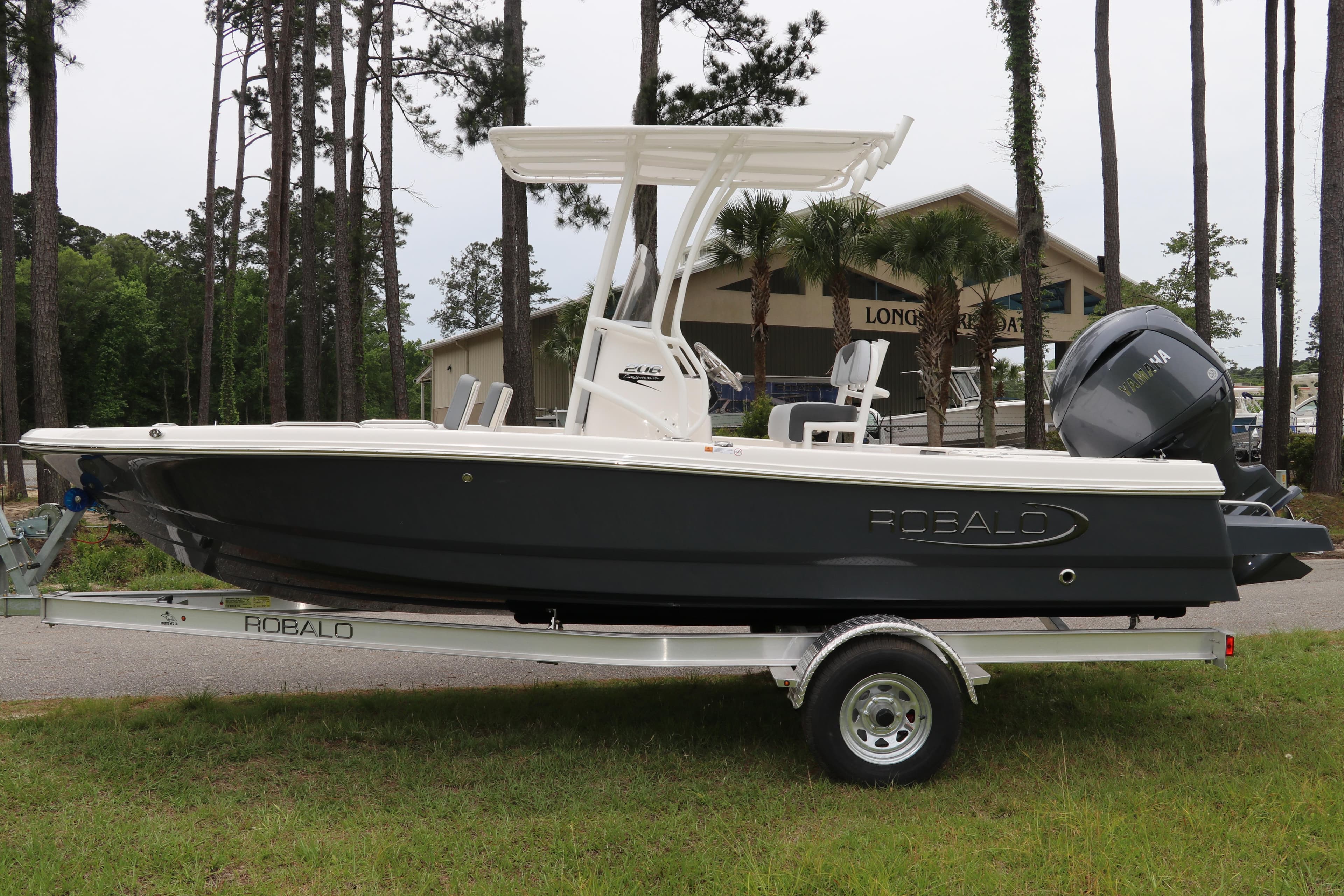 2026 Robalo 206 Cayman boat on trailer, parked outdoors near trees and building.