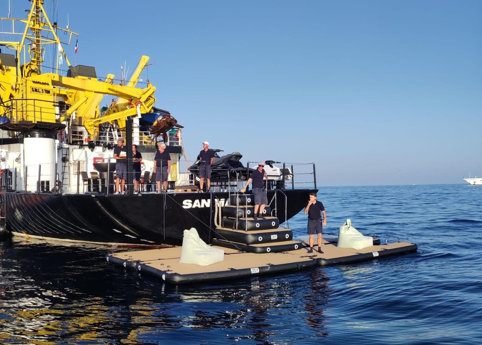People on a floating platform near a large ship, under clear blue skies.