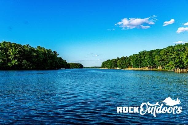 Scenic lake view with lush green trees under a clear blue sky, Rock Outdoors logo visible.