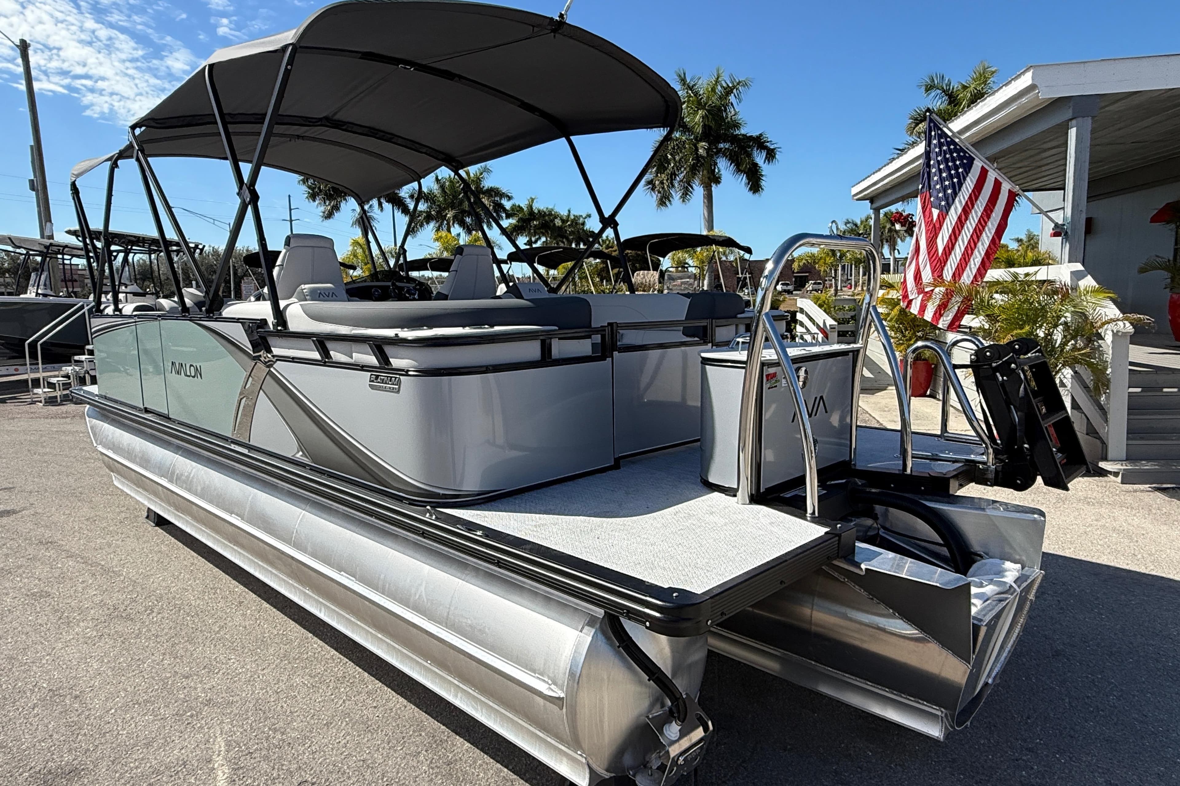 2026 Avalon 2385 LSZ Quad Lounger Tri Toon boat with American flag, parked outdoors.