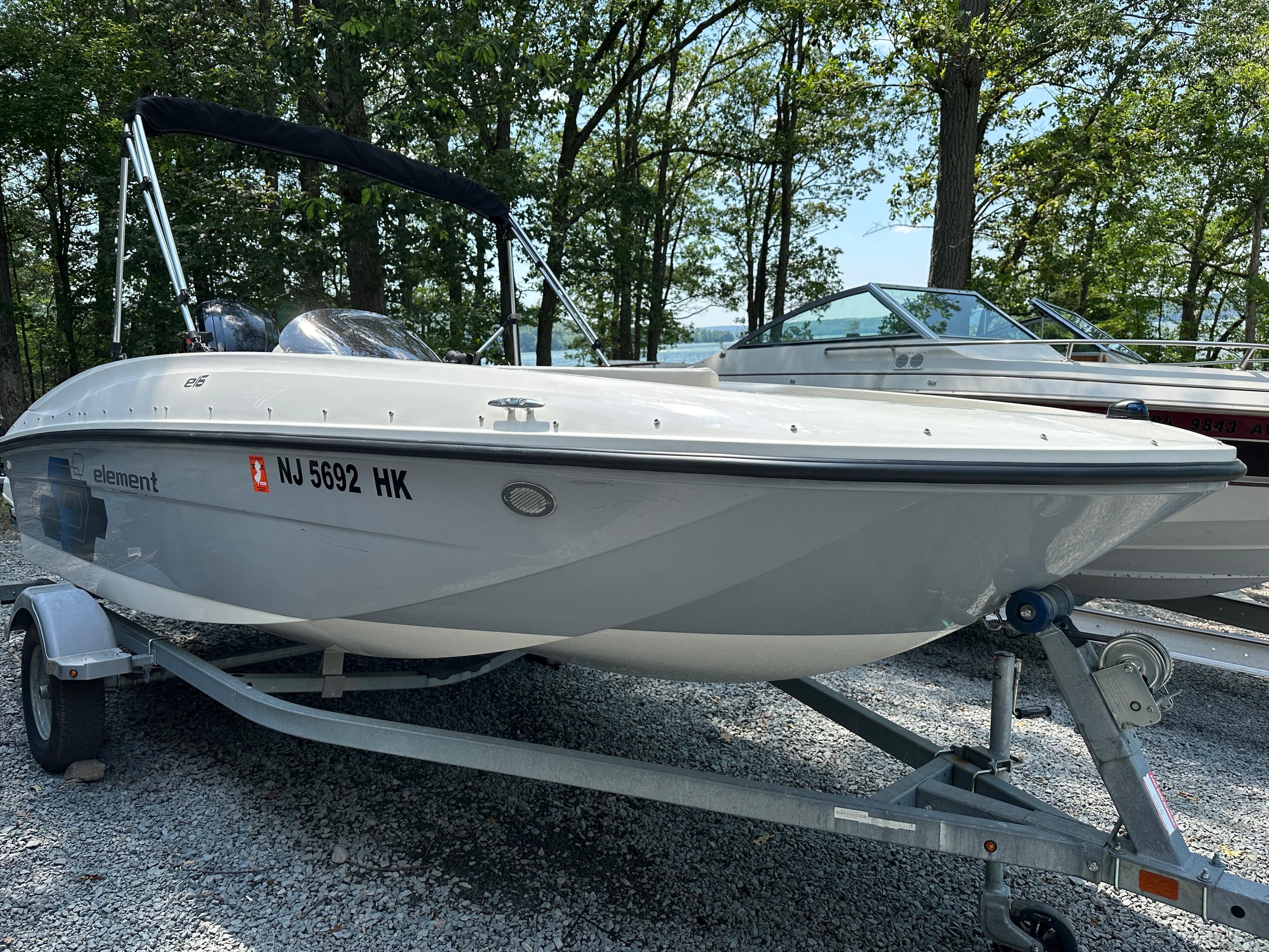 2019 Bayliner Element E16 boat on trailer, parked on gravel, surrounded by trees.