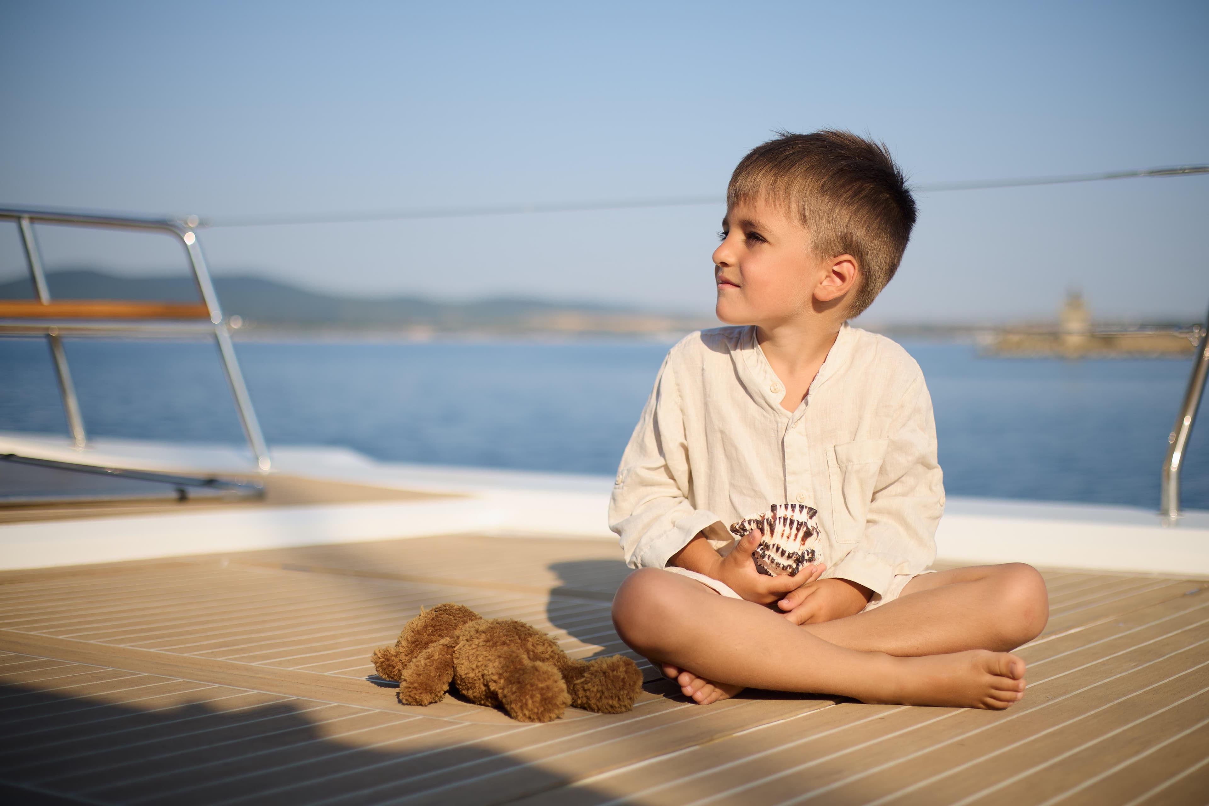 Young boy sitting on Omaya 50 Catamaran deck, holding a seashell, with a teddy bear nearby.
