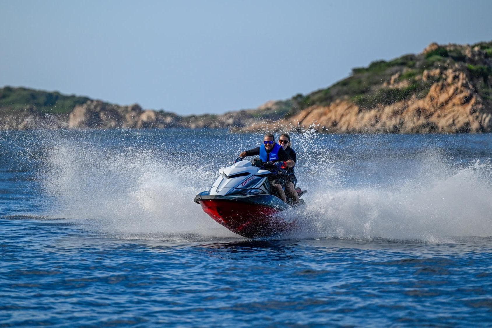 2024 Yamaha WaveRunner GP1800R SVHO speeding on water with two riders, scenic rocky background.