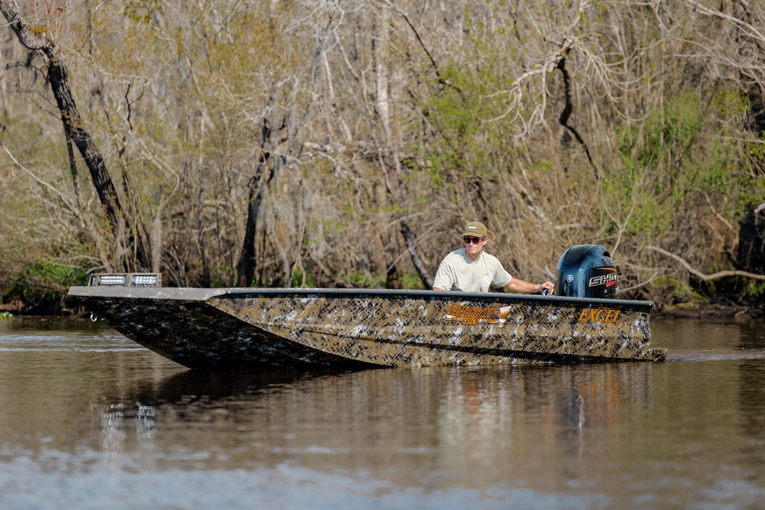 Man operating Excel 183 Tomahawk boat on a calm river, surrounded by trees.