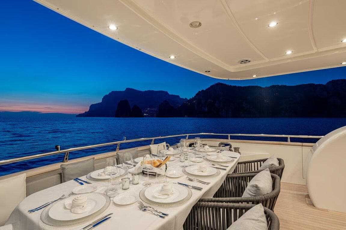 Luxurious dining setup on 2002 Maiora 24S yacht at sunset, overlooking serene ocean and cliffs.