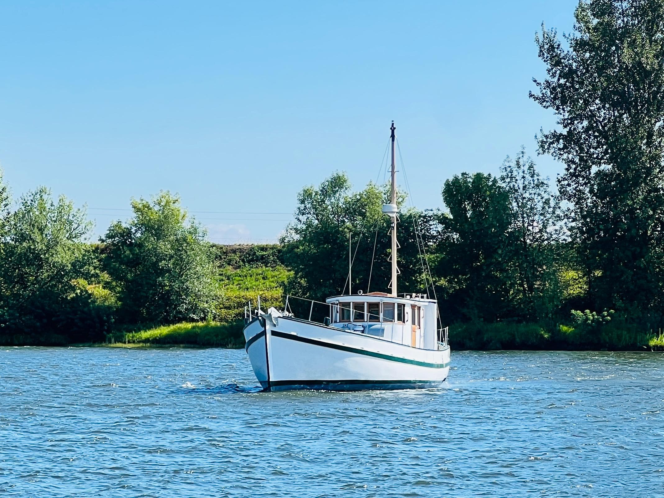 1955 Colin Archer 42 boat sailing on a calm river with lush green trees in the background.