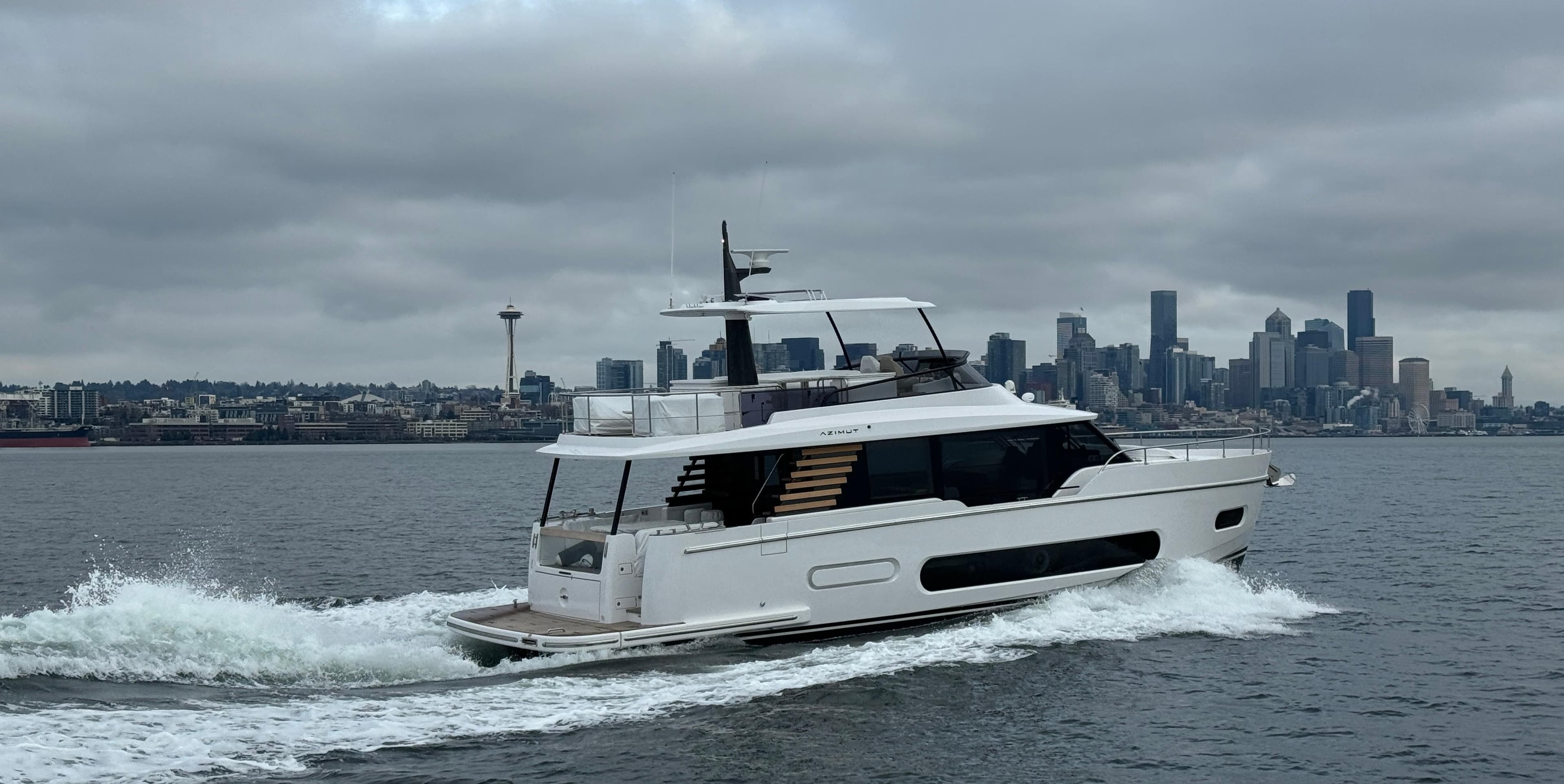 2025 Azimut Magellano 60 yacht cruising near a city skyline under cloudy skies.