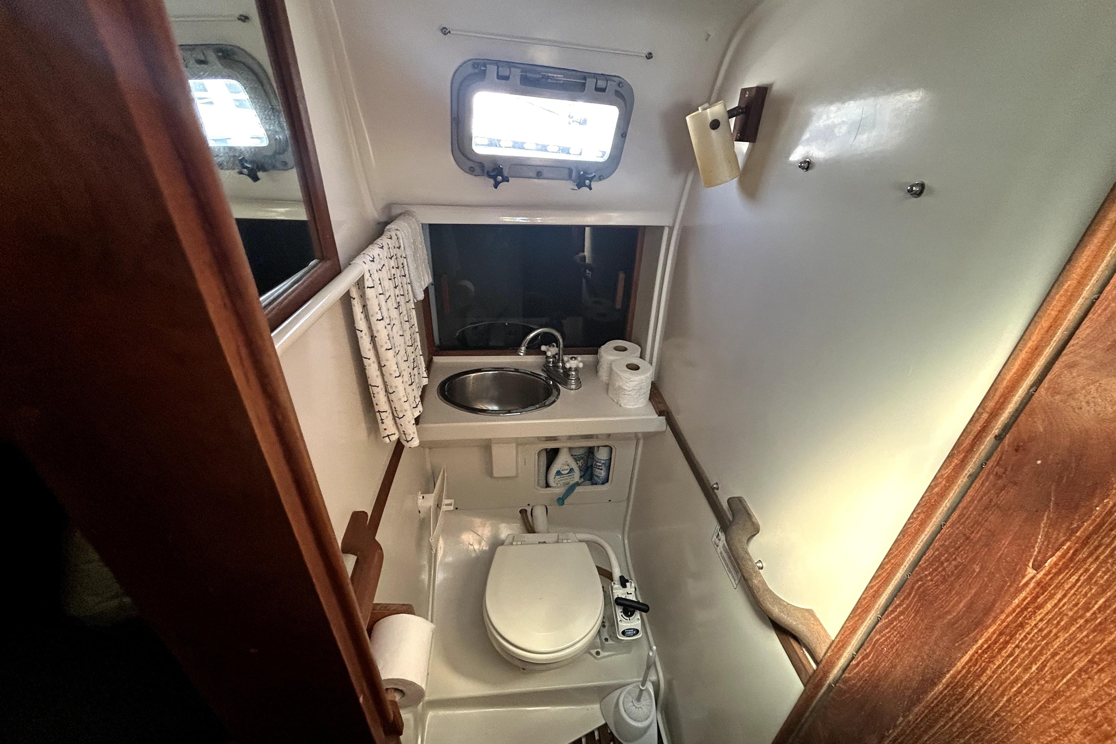 Compact bathroom in a 1981 Bayfield 32 sailboat with sink, toilet, and porthole window.
