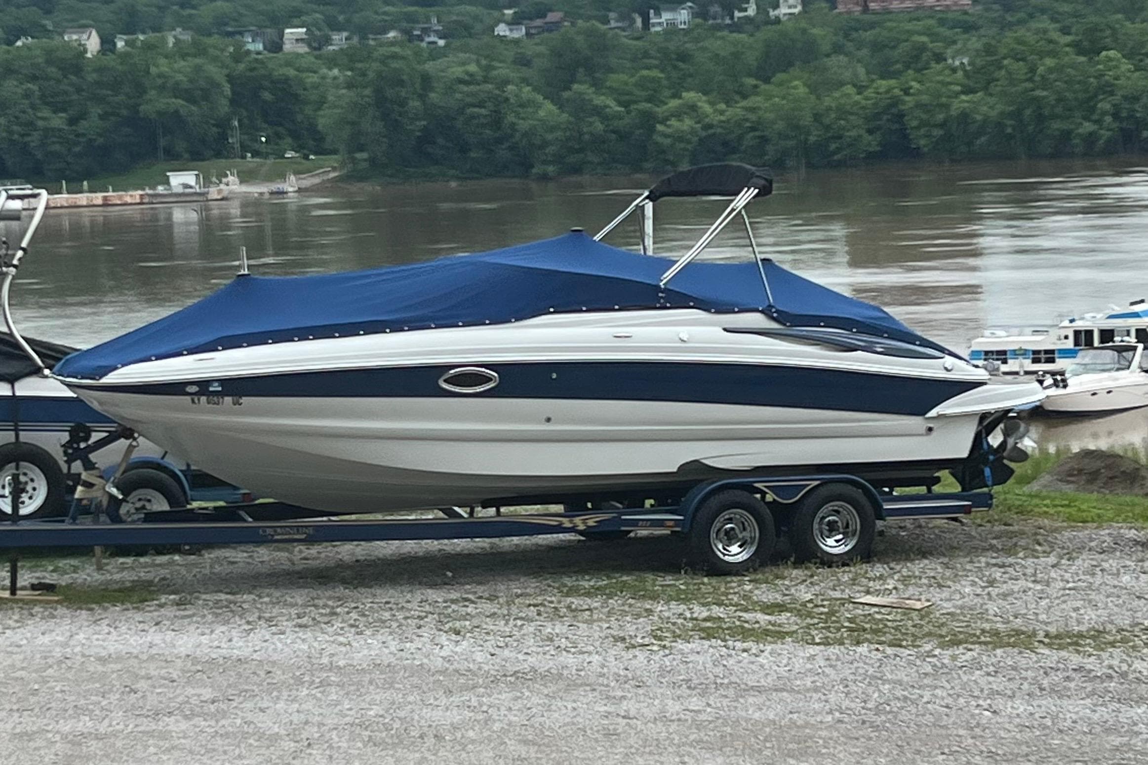 2006 Crownline 252 EX boat on trailer by riverbank, covered with blue tarp.