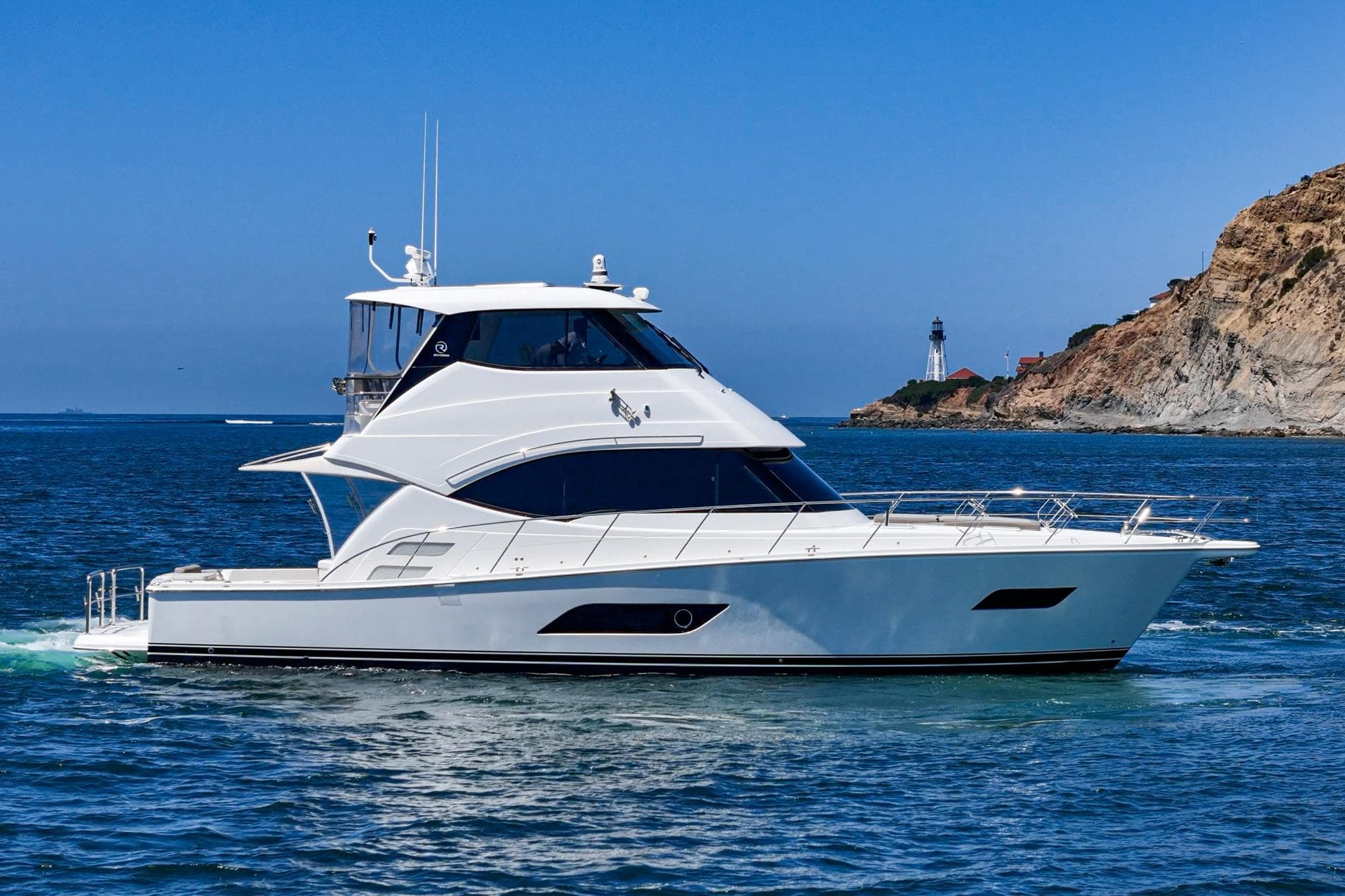 2019 Riviera 52 Enclosed Flybridge yacht cruising near a rocky coastline and lighthouse.