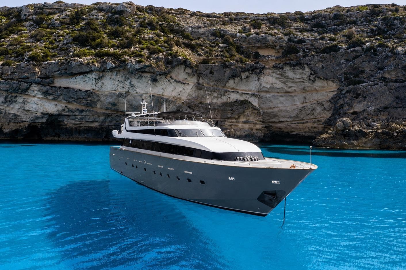 Luxurious 1995 Mondomarine 120 yacht anchored in clear blue waters near rocky cliffs.