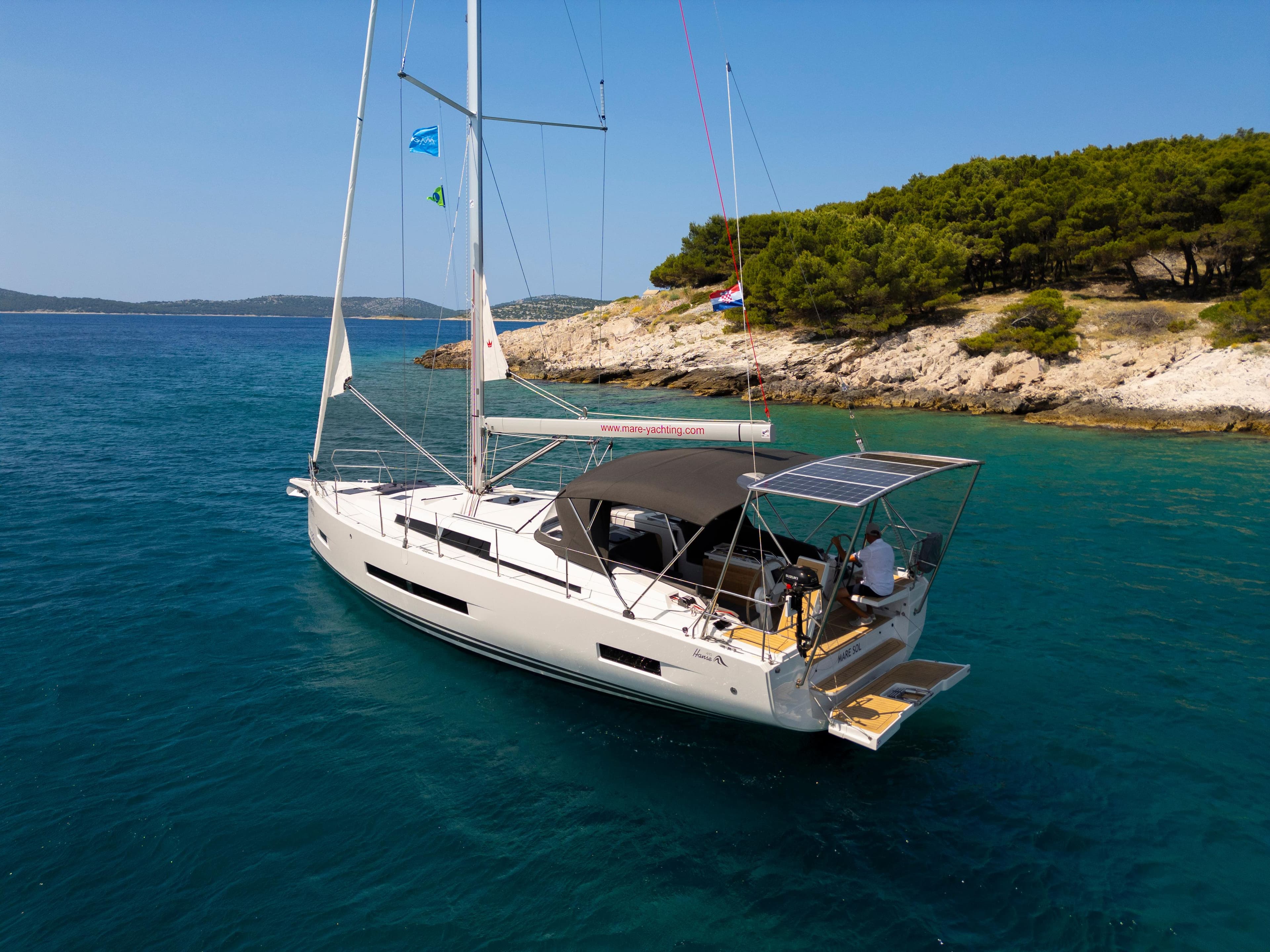 Sailing yacht Hanse 410, 2025 model, cruising in clear blue waters near a rocky coastline.