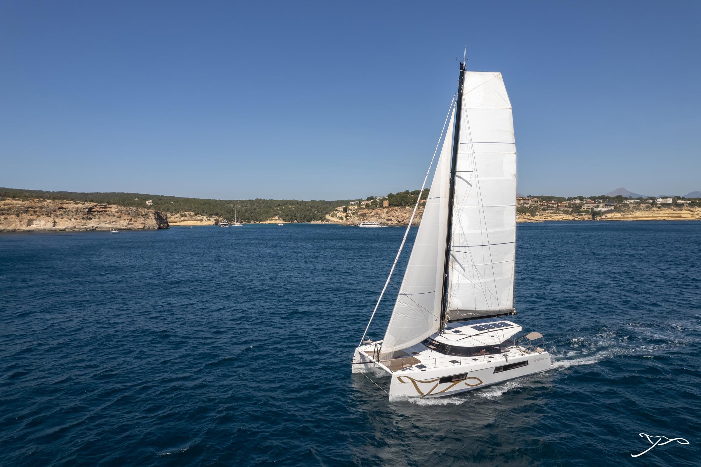 2025 Nautitech 44 Open catamaran sailing on clear blue waters near a rocky coastline.