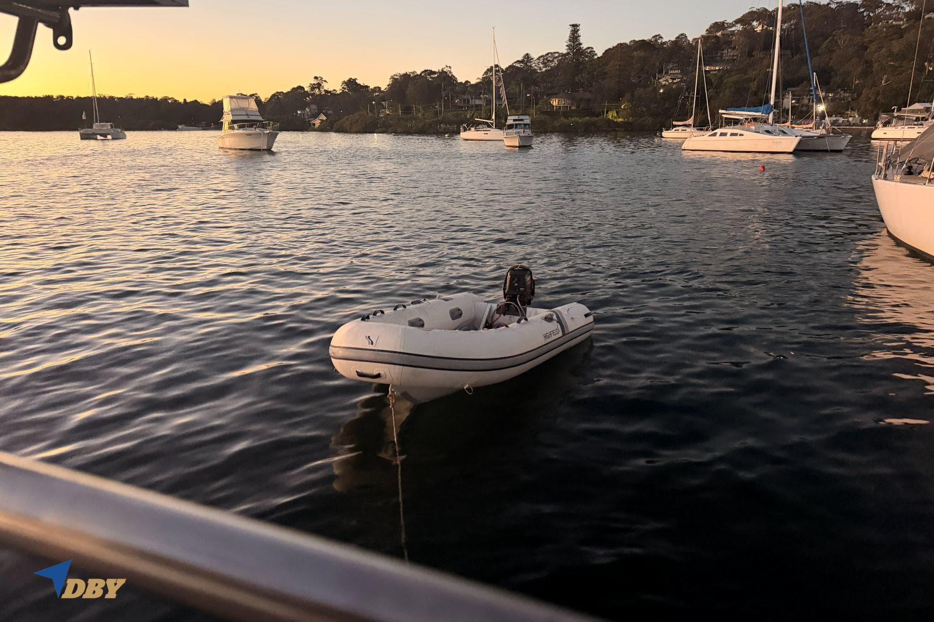 Inflatable boat on calm water at sunset with anchored sailboats nearby.