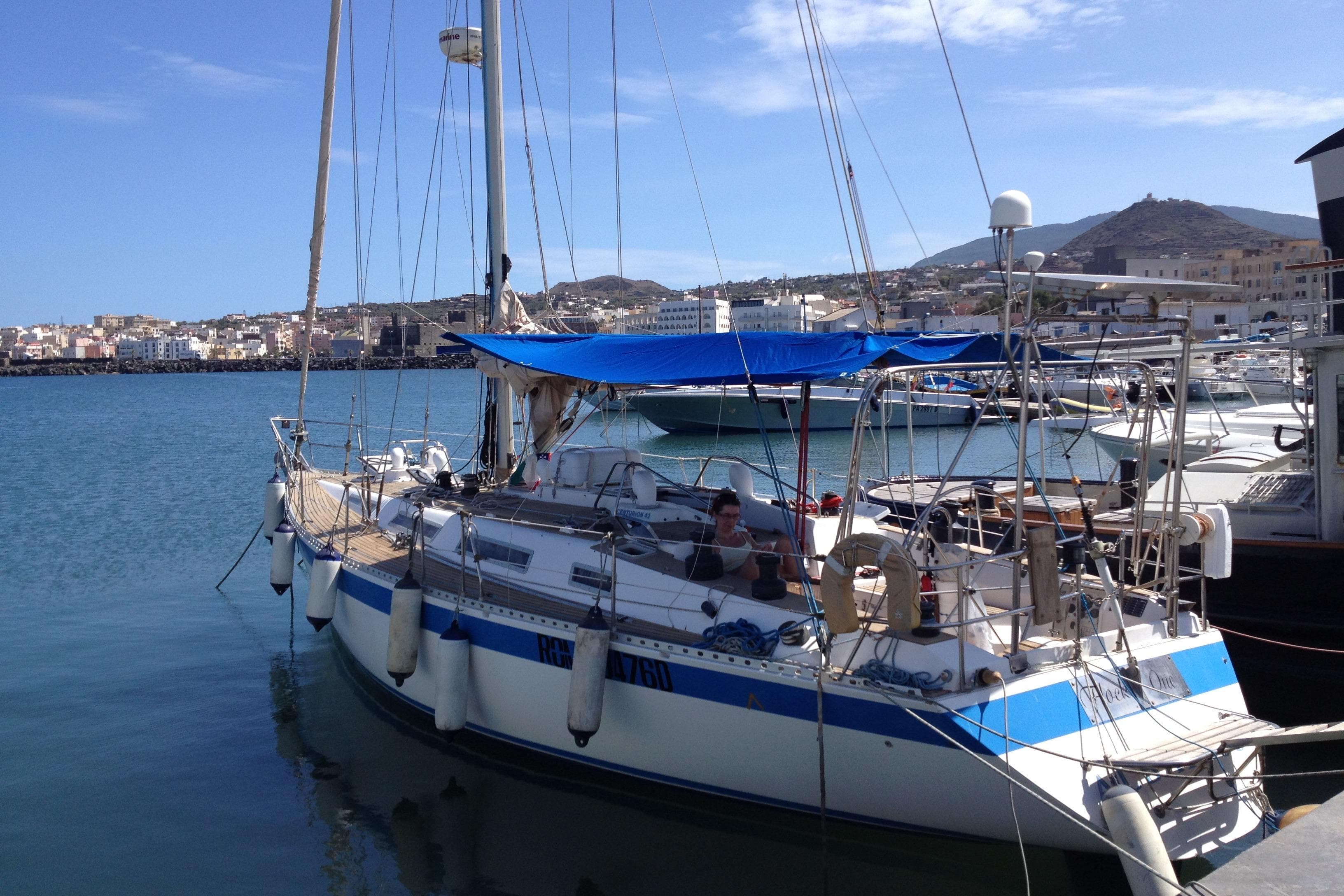 1987 Wauquiez Centurion sailboat docked in a sunny marina with cityscape background.