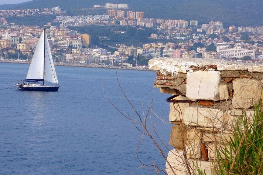 Sailboat on calm sea near coastal cityscape, with stone wall in foreground.